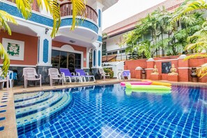 A swimming pool with lounge chairs and inflatable toys, surrounded by a building with a red and white facade and a balcony. - Baan Kanittha - 5 Bedrooms Pool Villa (Pattaya)