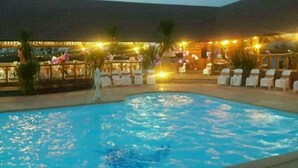 Een zwembad met een jacuzzi, omringd door ligbedden en tafels, en een overdekt gebied met lichtslingers.
