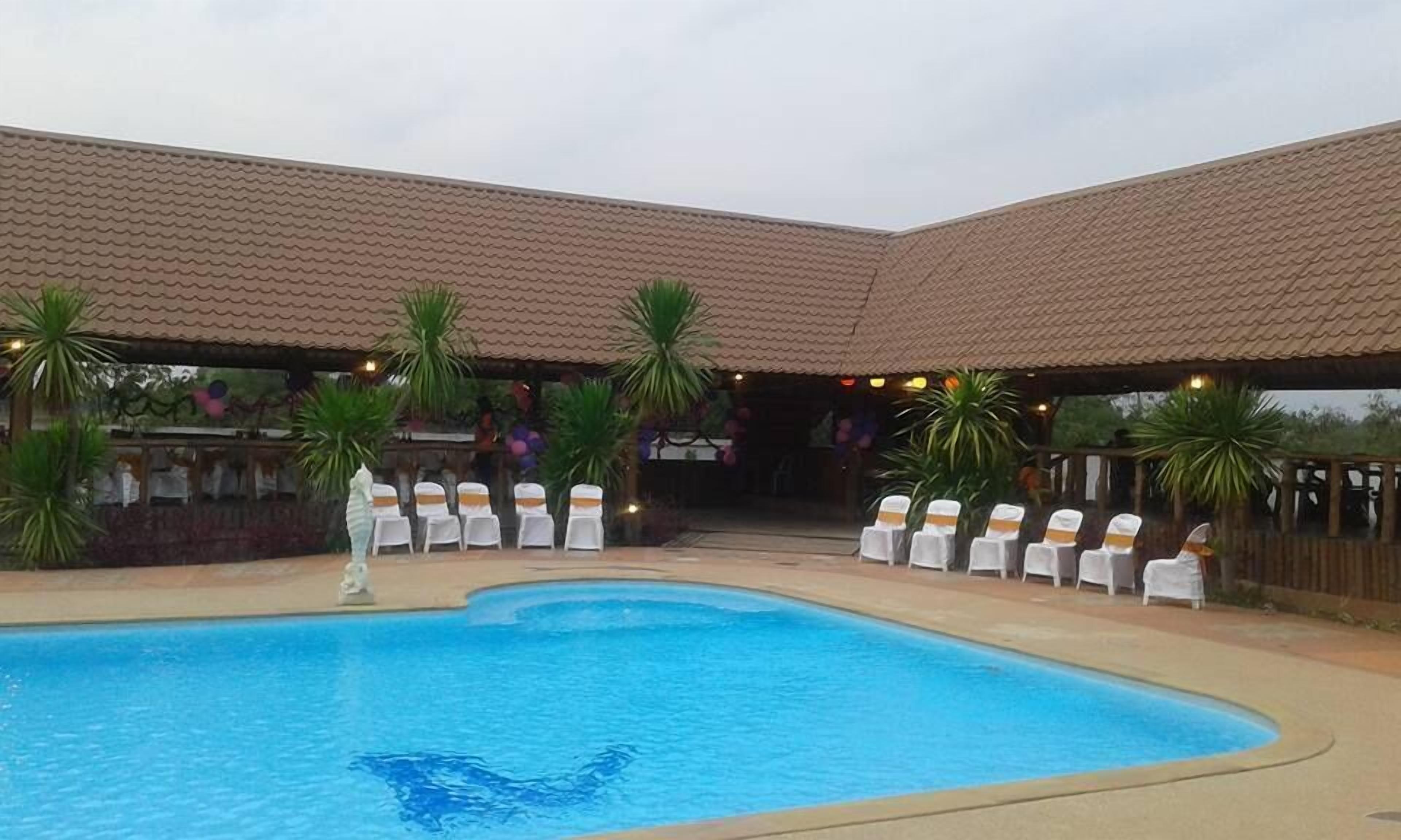 A swimming pool with chairs arranged around it, a covered area with a tiled roof, and palm trees.