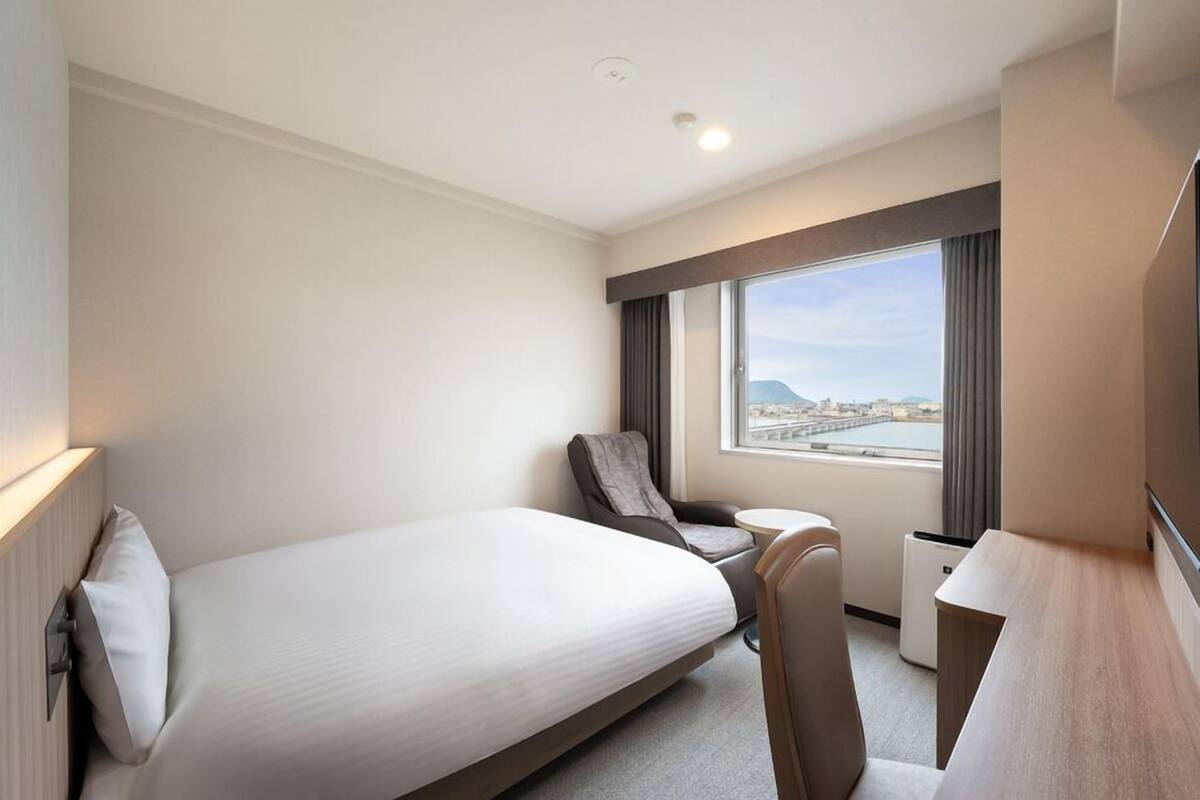 A hotel room with a bed, a desk, a chair, a small table, and a window with a view of a cityscape.