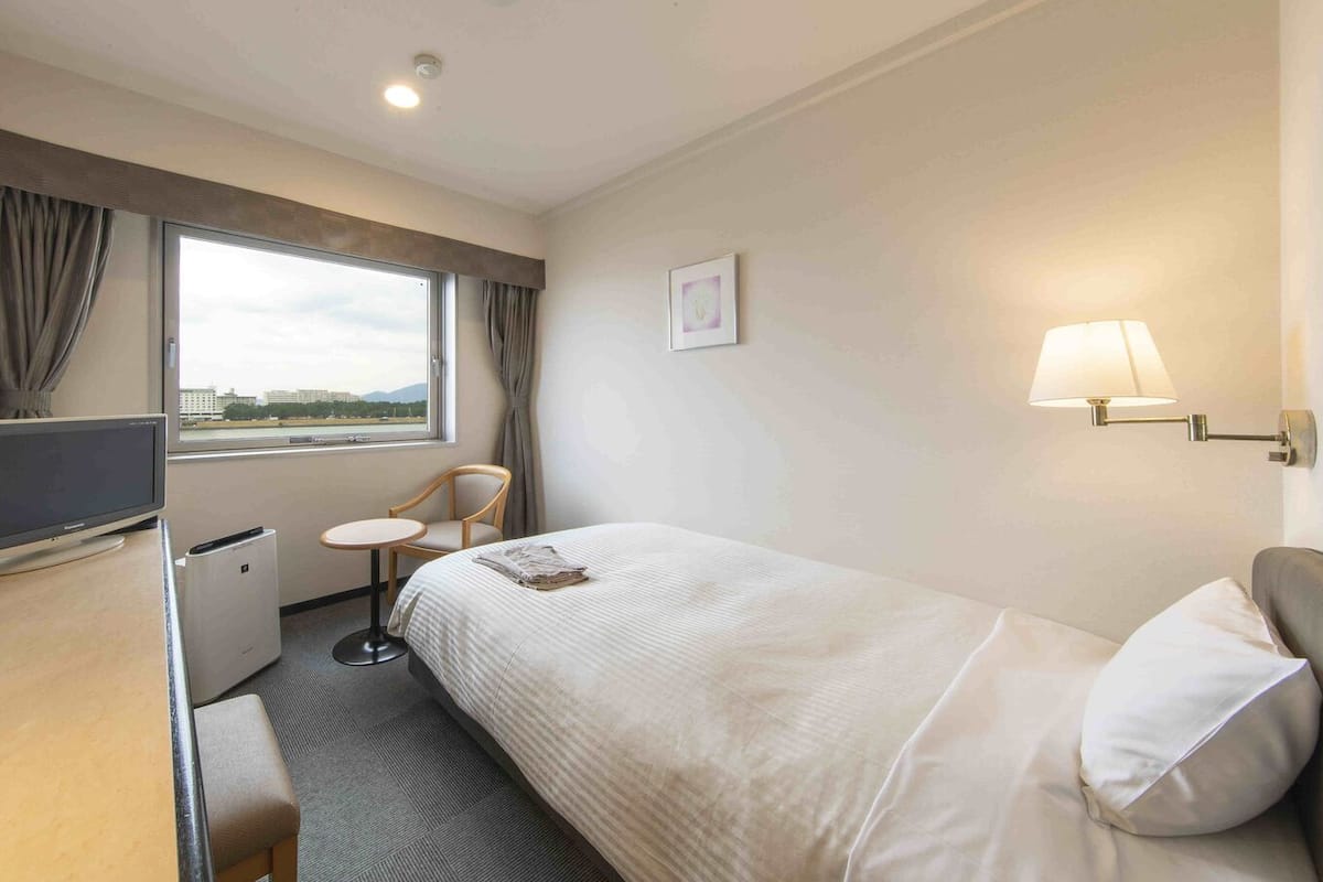 A hotel room with a bed, a desk with a computer, a chair, a small table, and a window with curtains.