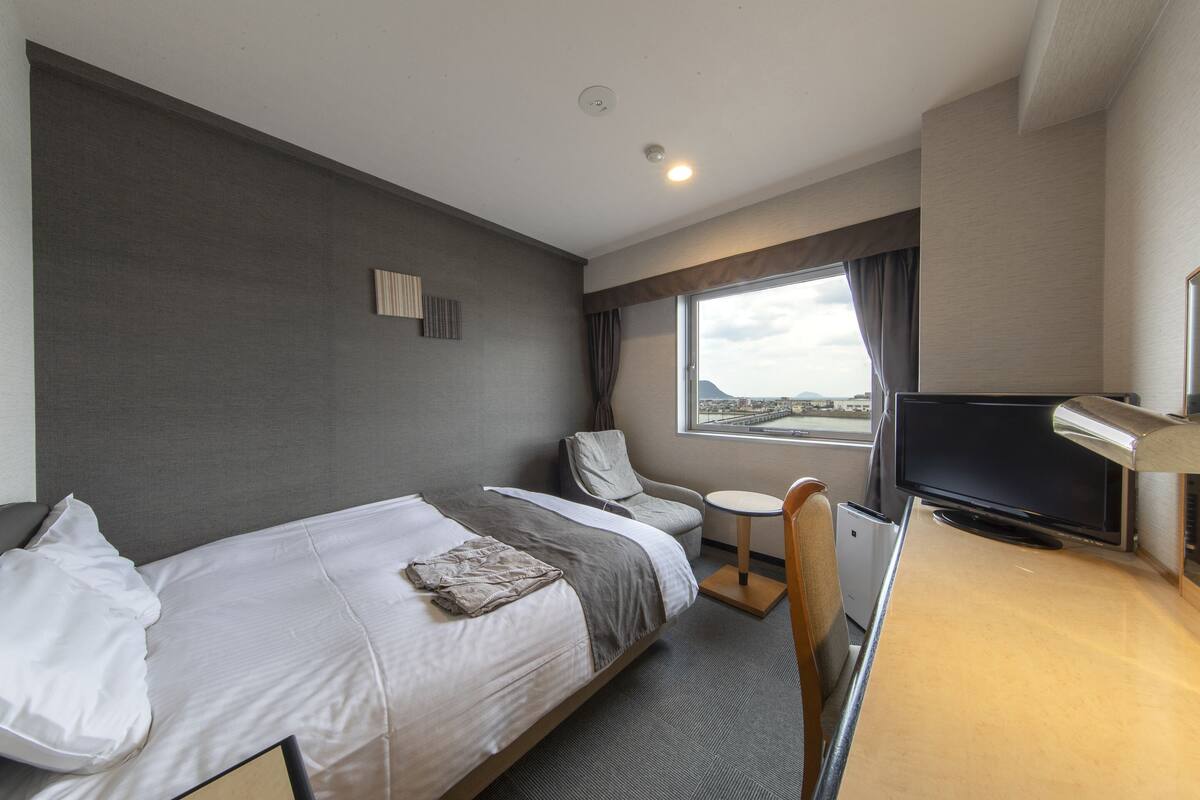 A hotel room with a bed, a chair, a small table, a TV, and a window with a view.