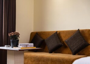 Una habitación con un sofá marrón, dos cojines oscuros, una mesita con una planta en maceta y libros.
