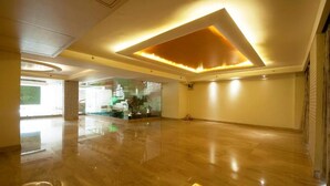 A spacious room with a polished wooden floor, a modern ceiling design with recessed lighting, and a glass partition with a view of a staircase and greenery. - Mayur Hotel (New Delhi)