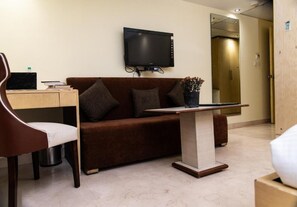 A hotel room with a sofa, a desk with a laptop, a television, and a small table with a plant. - Mayur Hotel (New Delhi)