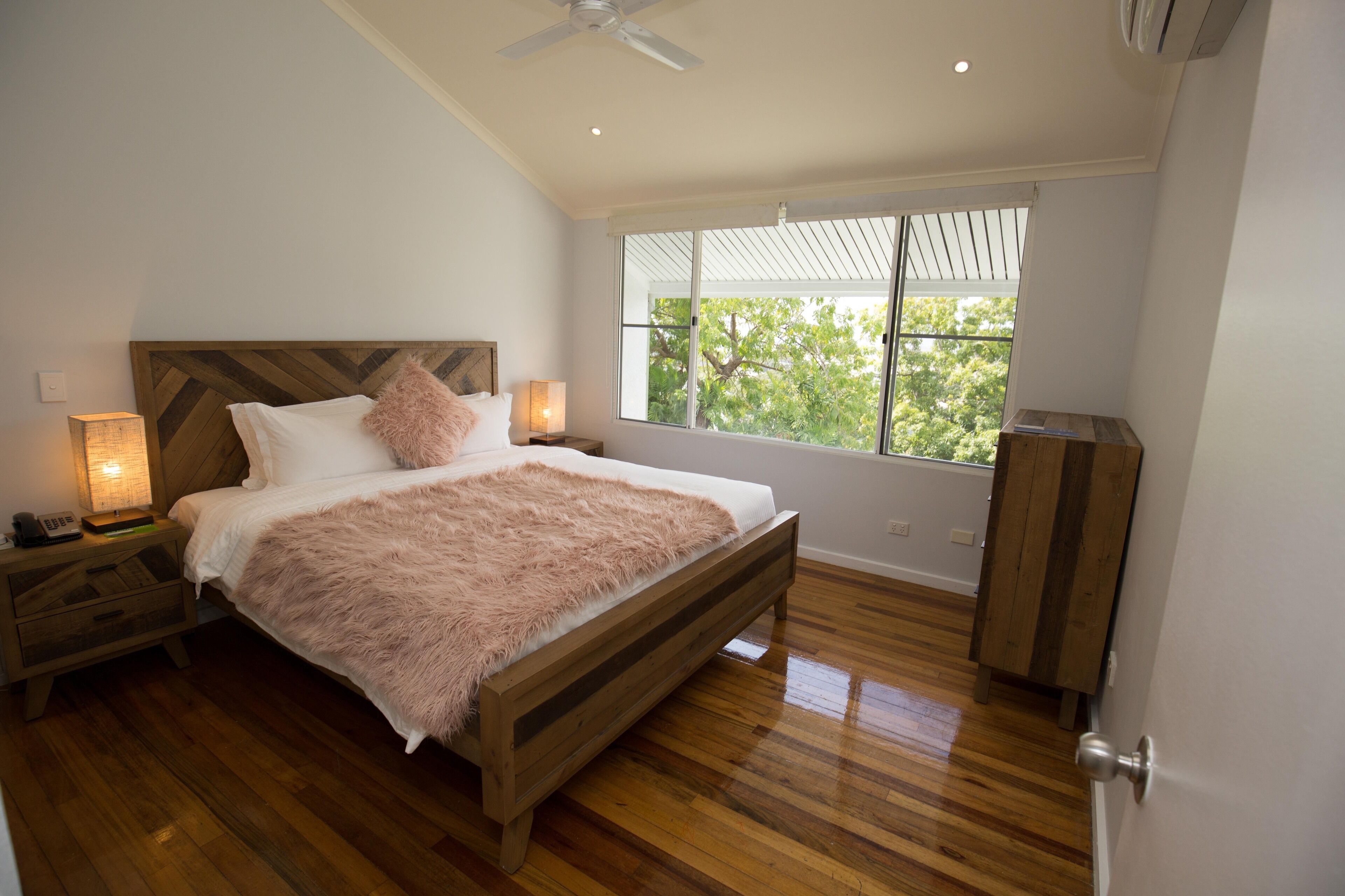 A bedroom with a wooden bed, two bedside lamps, a nightstand, and a window with a view of trees.