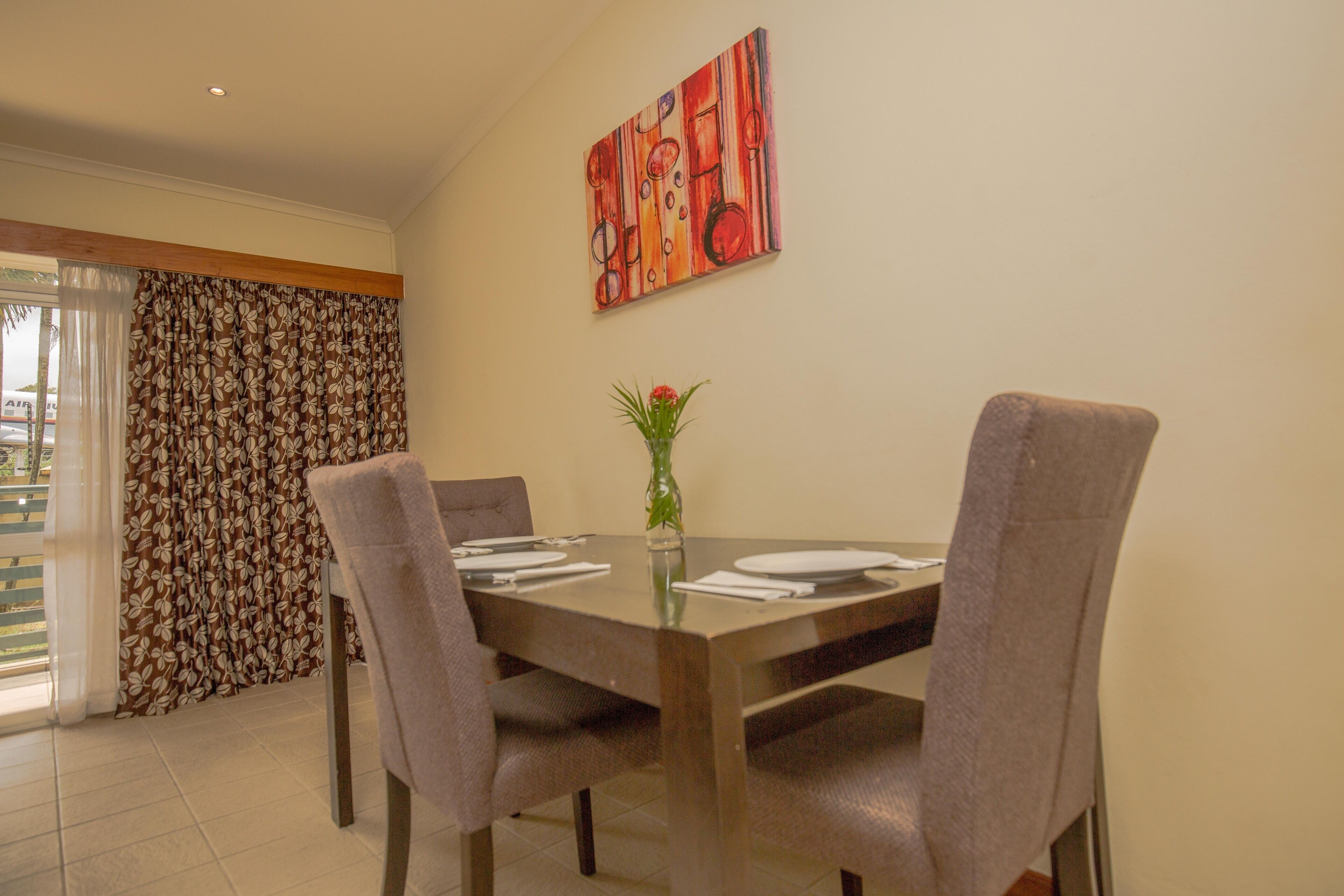 A dining area with a table set for four, a vase with flowers, and a painting on the wall.