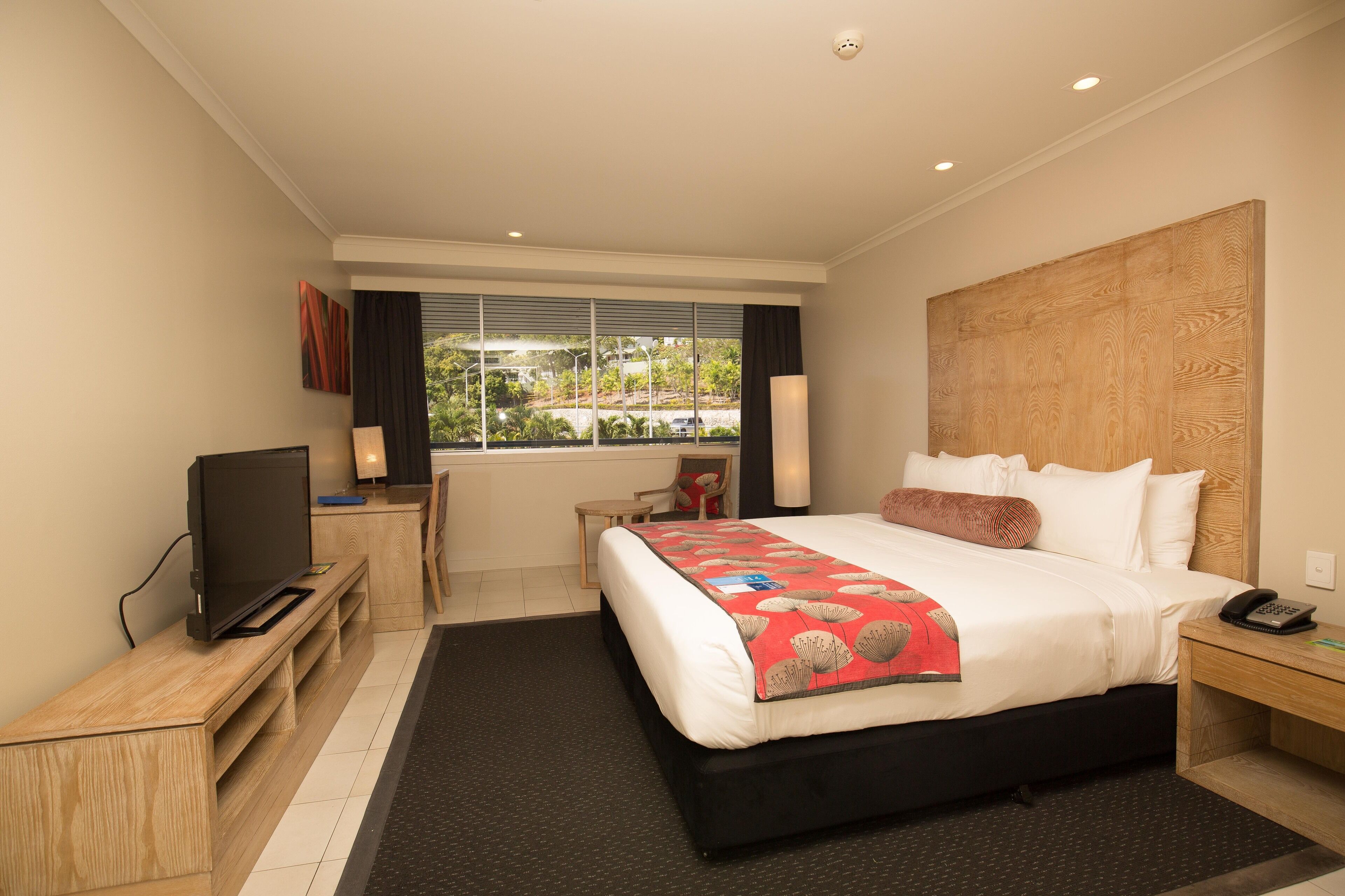 A modern hotel room with a large bed, a desk with a flat-screen TV, a chair, and a window with a view of greenery.