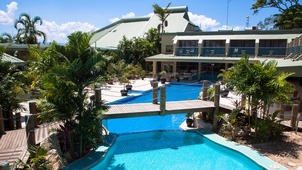 A large outdoor swimming pool with multiple lanes, surrounded by palm trees and a building with a green roof. - Gateway Hotel (Port Moresby)