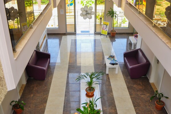 Un vestíbulo con una escalera central, dos sofás morados, una mesa blanca y plantas en macetas.