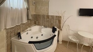 A modern bathroom with a freestanding bathtub, stone wall, a small table with chairs, and a flat-screen TV mounted on the wall. - Antica Mateola Rooms&Suites (Matera)