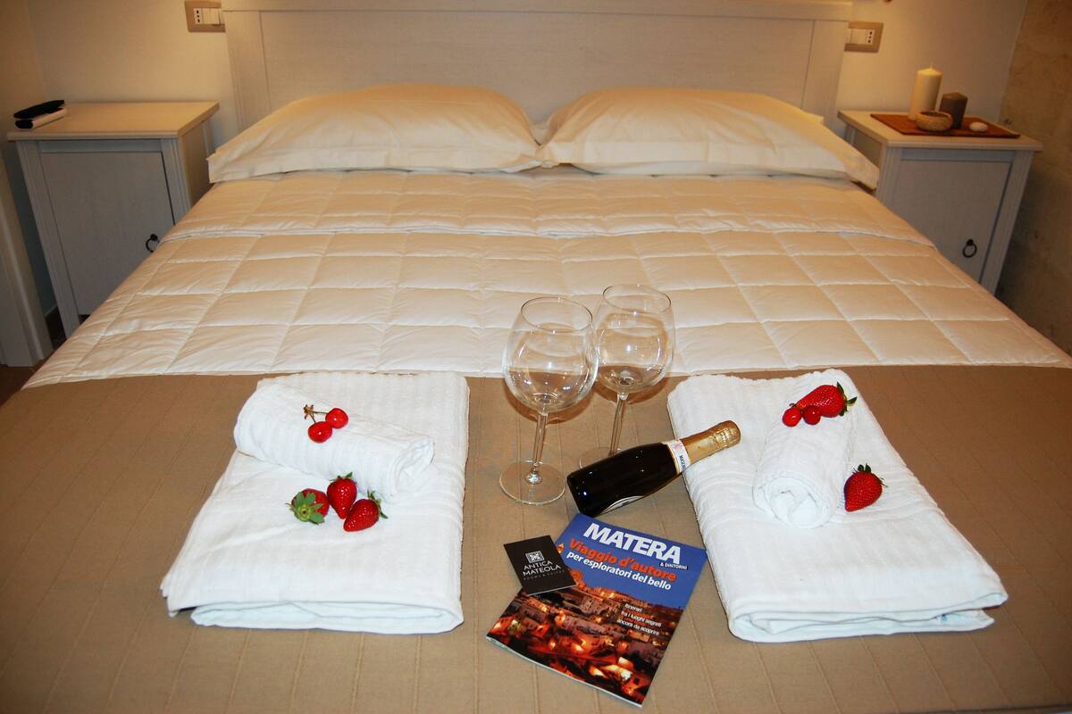 Een net opgemaakt bed met witte lakens, twee lege wijnglazen, een fles champagne en een brochure met de titel 