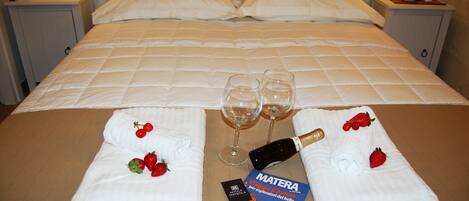 A neatly made bed with white linens, two empty wine glasses, a bottle of champagne, and a brochure titled "MATERA" on the bed.