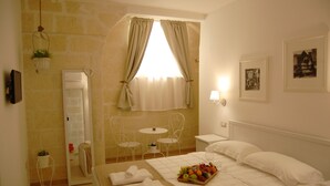 A hotel room with a bed, a bedside table with a fruit basket, a small table with chairs, a mirror, and a window with curtains. - Antica Mateola Rooms&Suites (Matera)