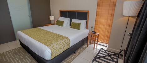 A hotel room with a bed, bedside table, lamp, and a window with blinds.