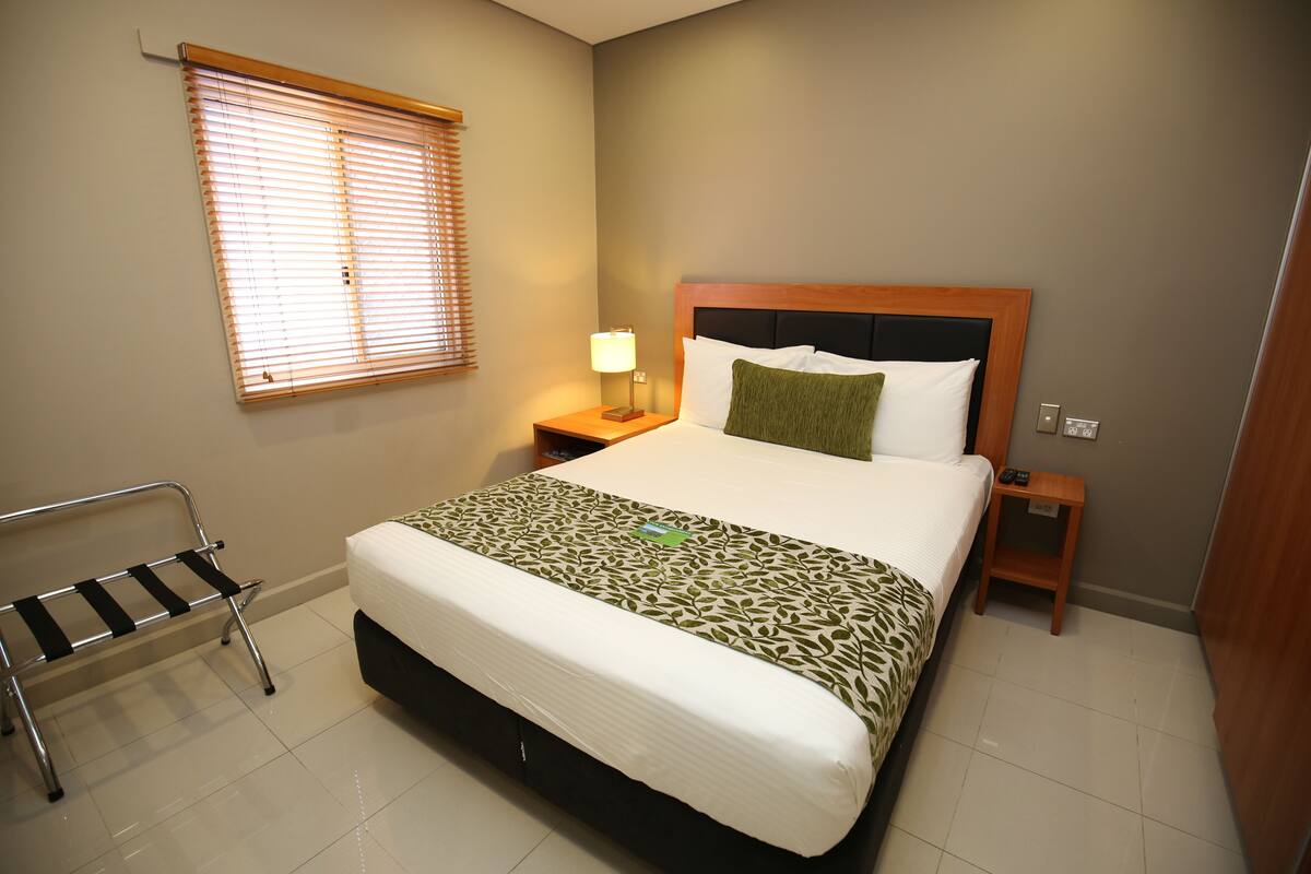 A hotel room with a large bed, a bedside table with a lamp, a bench, and a window with blinds.