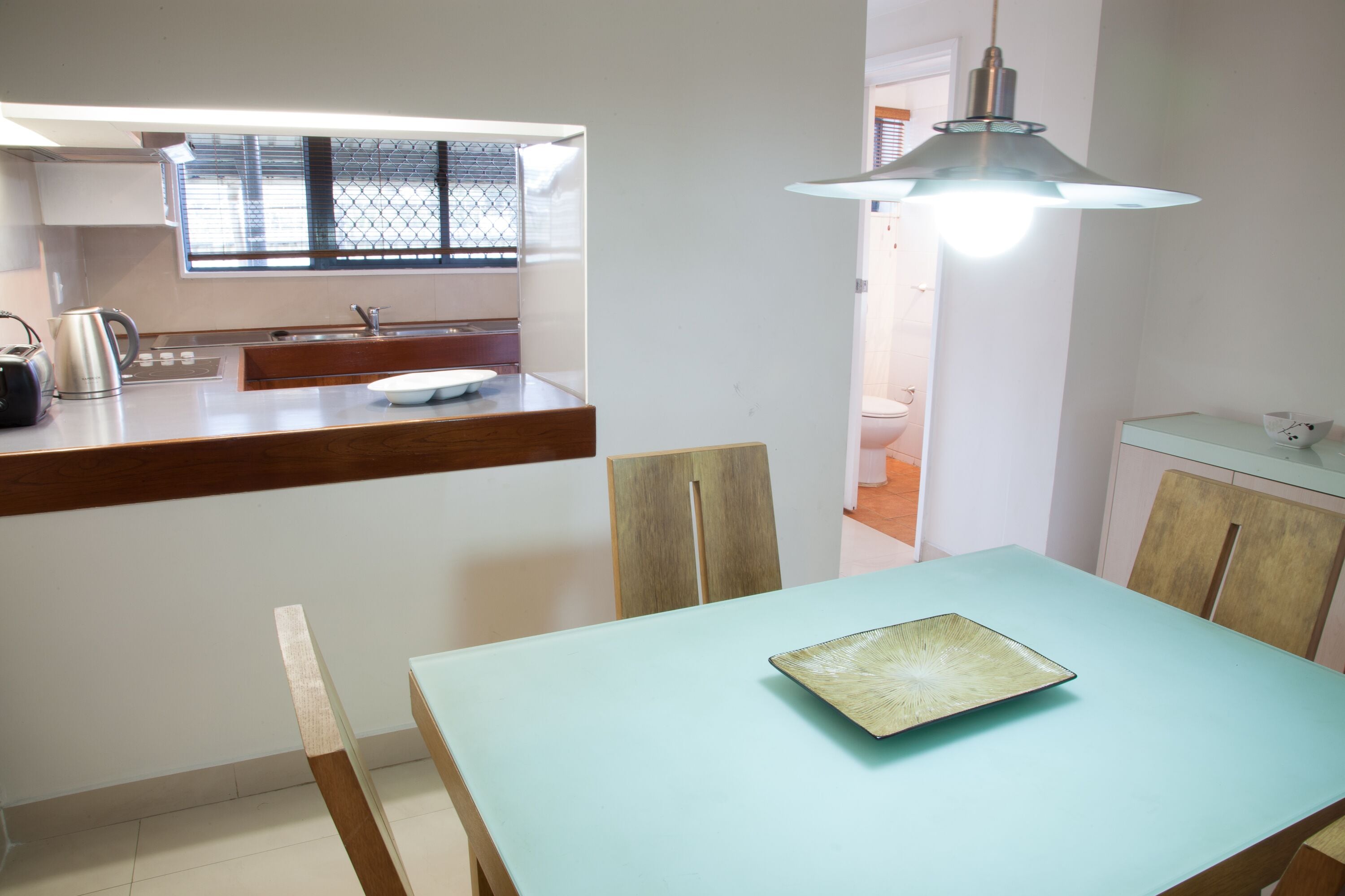 Un espace repas comprenant une table, des chaises, un luminaire, ainsi qu’un coin cuisine avec une fenêtre et un comptoir.