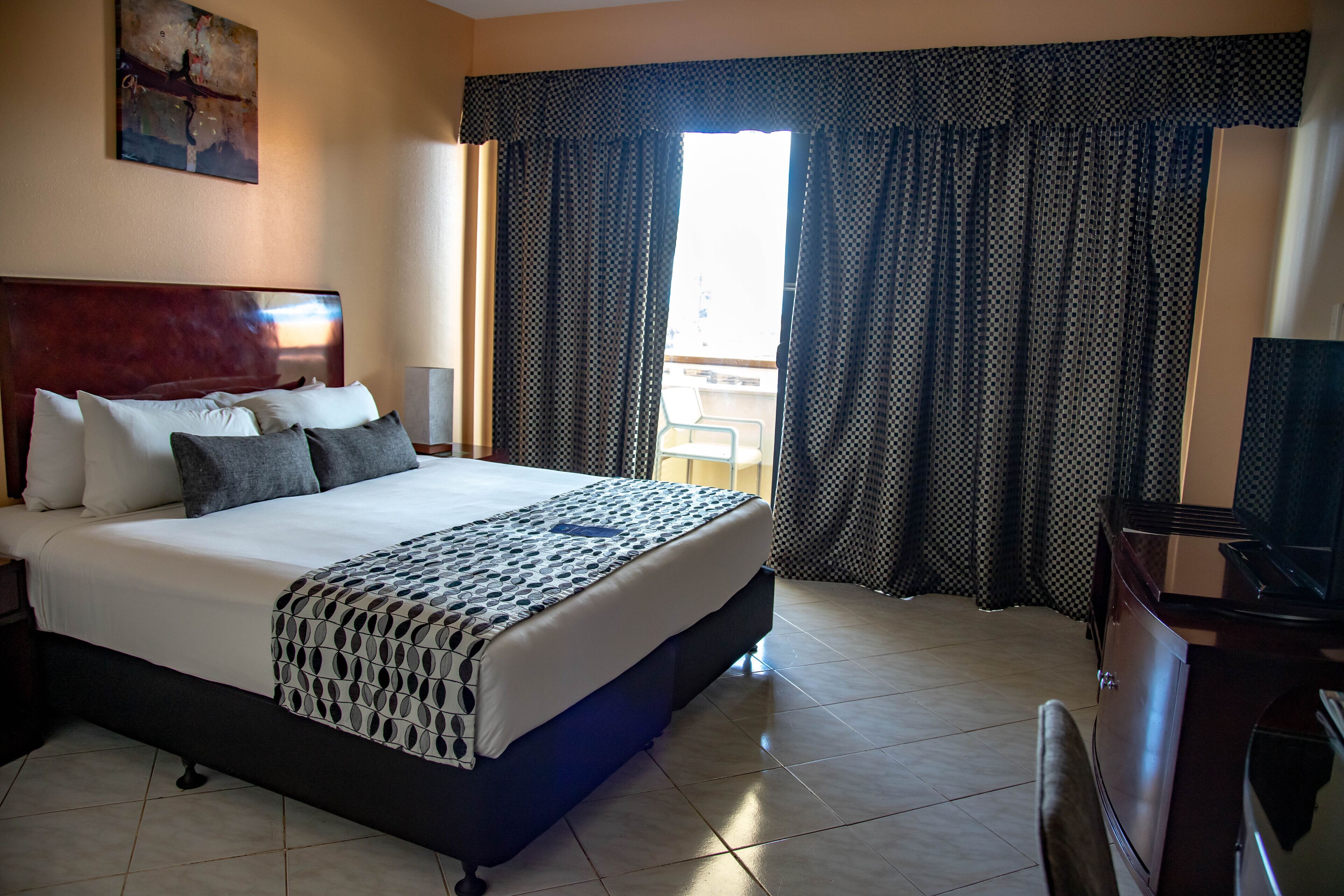A hotel room with a large bed, a wooden headboard, a flat-screen TV, and a balcony with curtains.