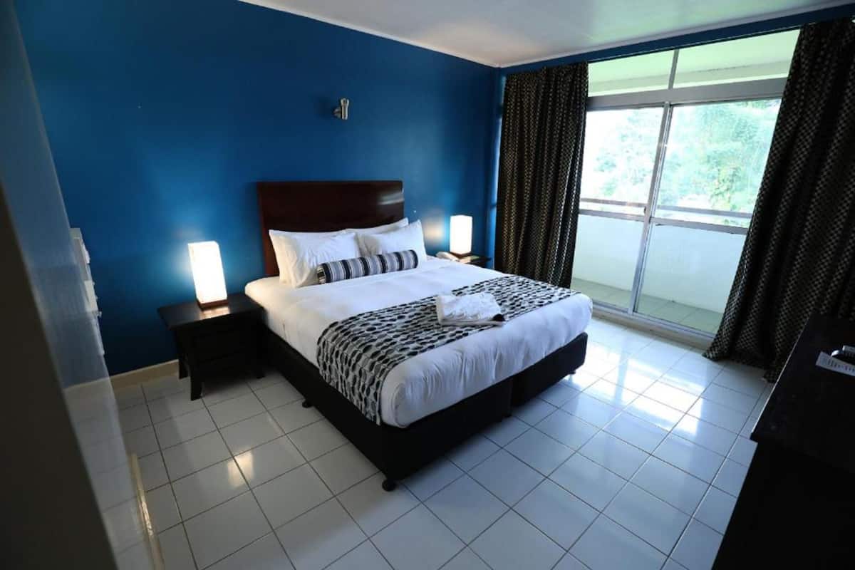 Une chambre d’hôtel avec un grand lit, deux tables de chevet, une fenêtre avec des rideaux et une vue sur la verdure.
