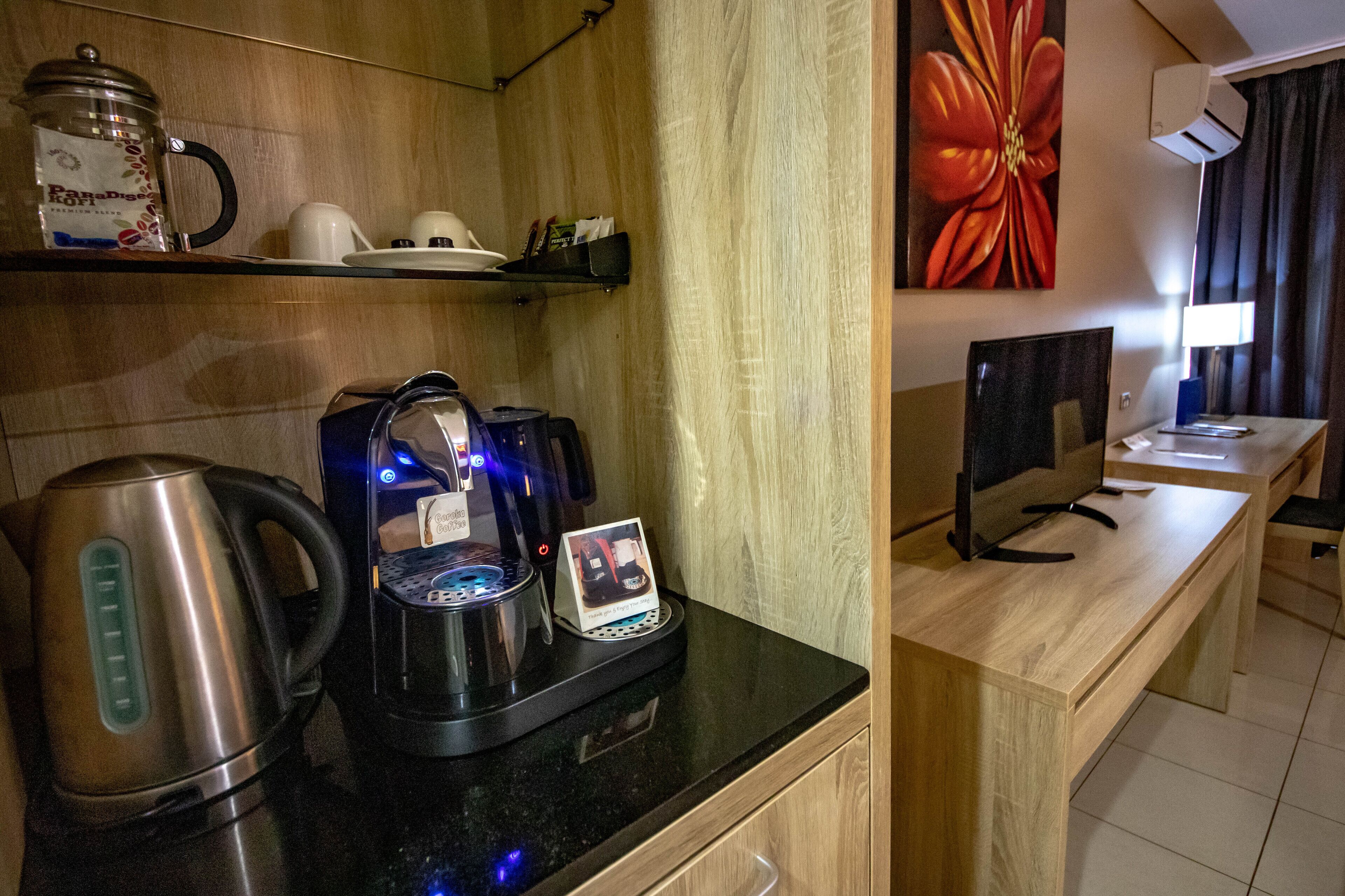 A kitchenette with a coffee maker, kettle, and a digital coffee pot. There is a computer on a desk and a painting on the wall.