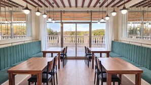 Un comedor con mesas de madera y bancos tapizados en verde, ventanas amplias con vista a un balcón e iluminación colgante.
