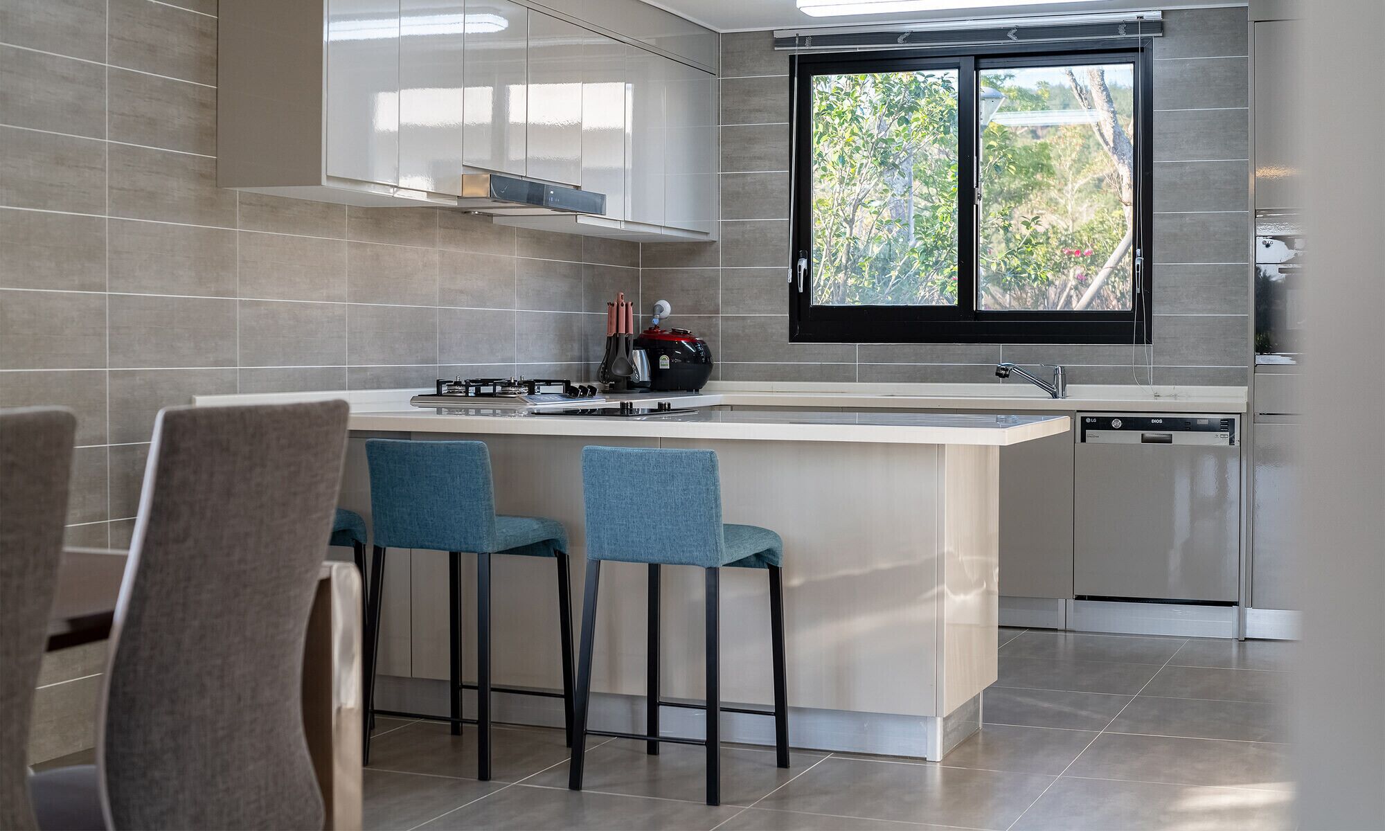Una cocina moderna con una isla grande, dos taburetes de bar, lavavajillas y una ventana con vistas al verdor.