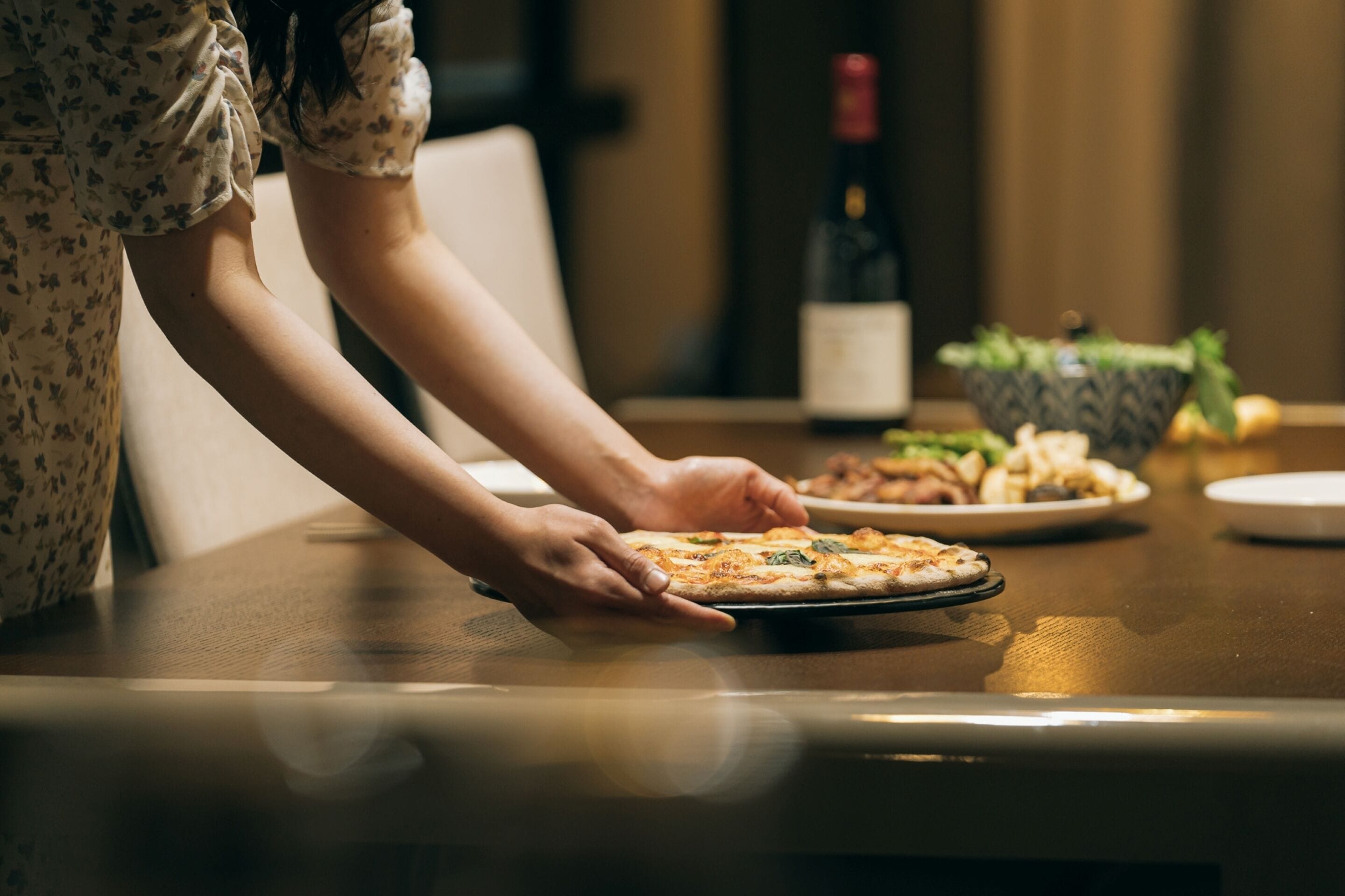 Iemand legt een pizza op een tafel, met een saladeschaal en een fles wijn op de achtergrond.
