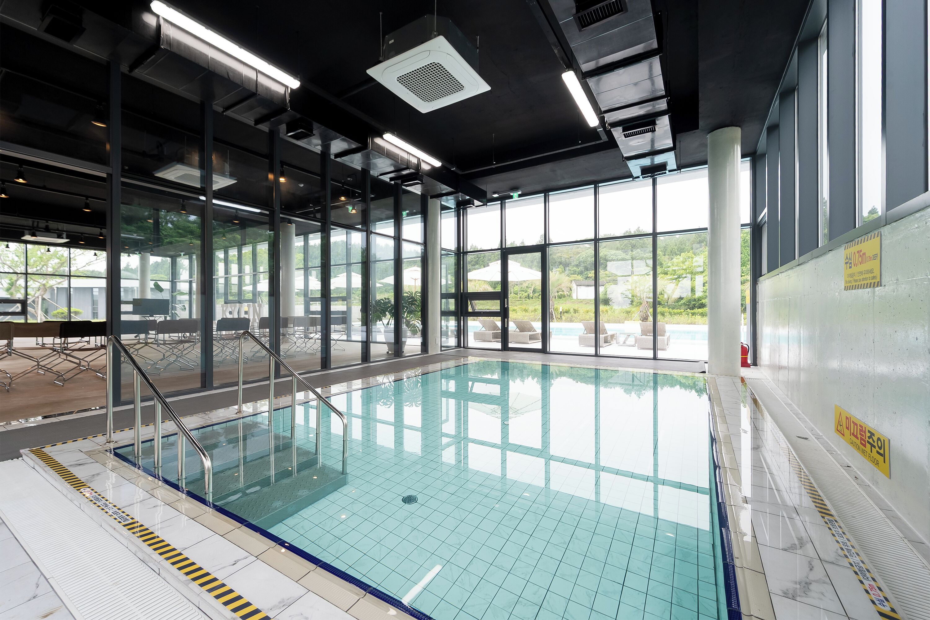 Una piscina interior con paredes de vidrio transparente, suelo de baldosas y escalera de metal.