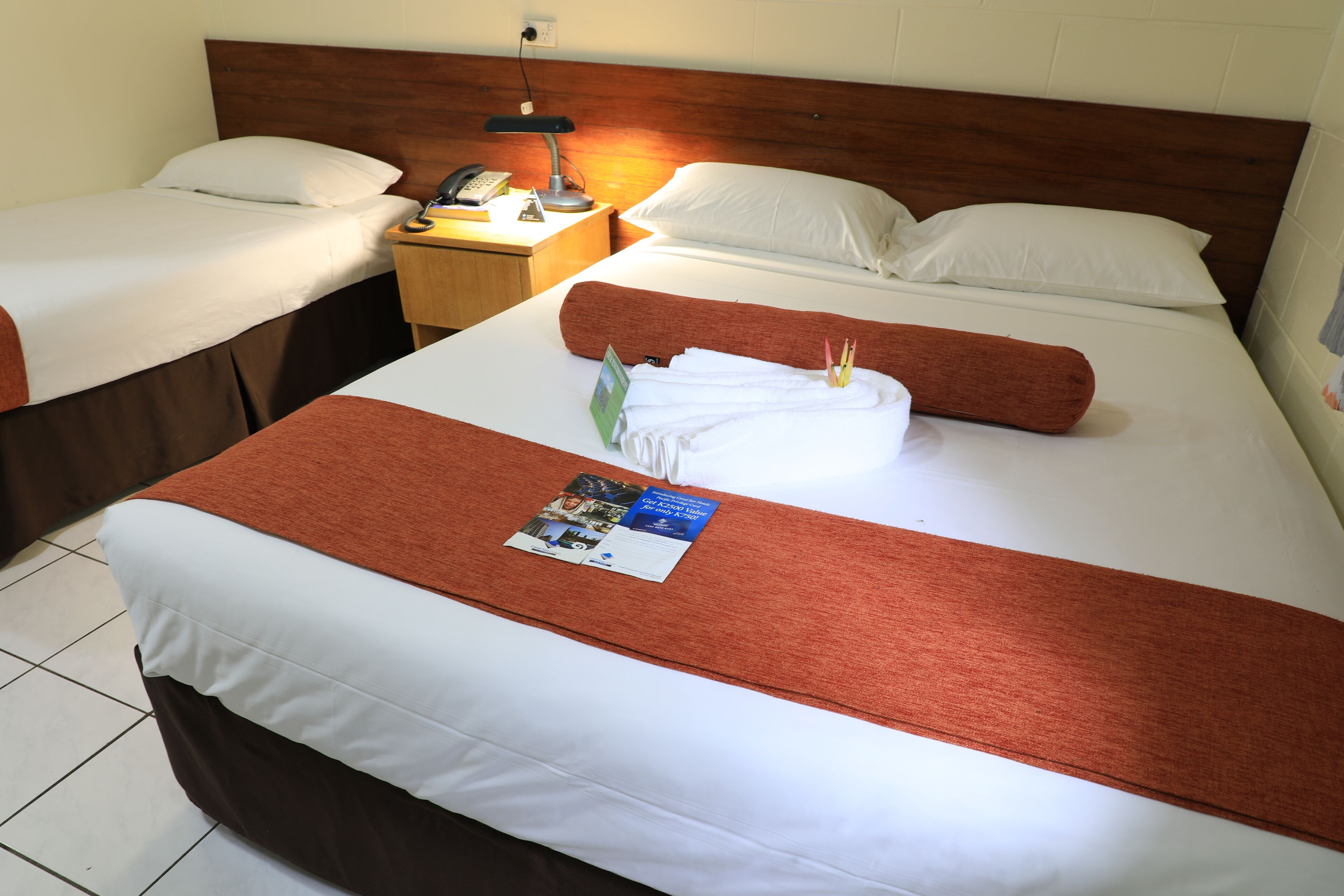 Een hotelkamer met twee bedden, een nachtkastje met een lamp, een telefoon en een brochure op de bedden.