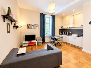 A modern living room with a grey sofa, a flat-screen TV, a dining table with chairs, a small kitchen area with a microwave, and a window with curtains. - Mosaic Center Apartments (Riga)