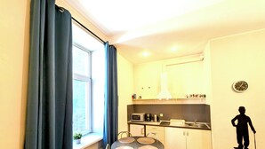 A compact kitchen with a dining table and chairs, a window with curtains, and a wall-mounted clock. - Mosaic Center Apartments (Riga)