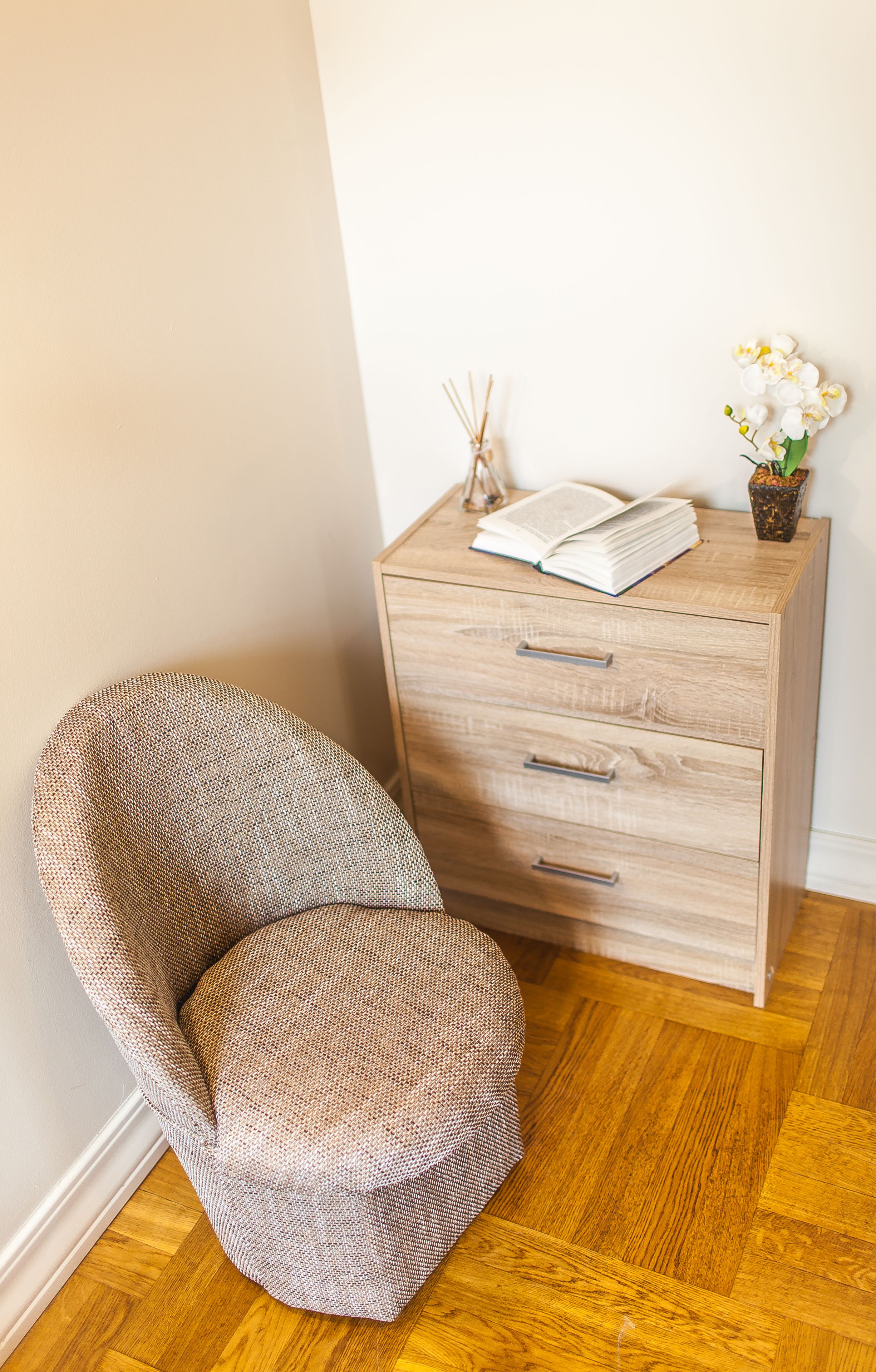 Une pièce d’angle avec un meuble en bois, une chaise en osier et une petite table sur laquelle se trouvent un livre et un vase de fleurs.