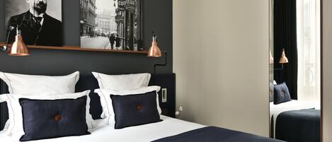 Una cama bien hecha con ropa de cama blanca y azul marino, dos lámparas fijadas a la pared y un cuadro colgado sobre la cama.