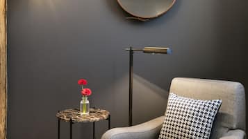 Une pièce moderne avec un mur gris, un miroir rond, un fauteuil gris agrémenté d’un coussin à motifs, une petite table ronde sur laquelle est posé un vase de fleurs et un lampadaire.