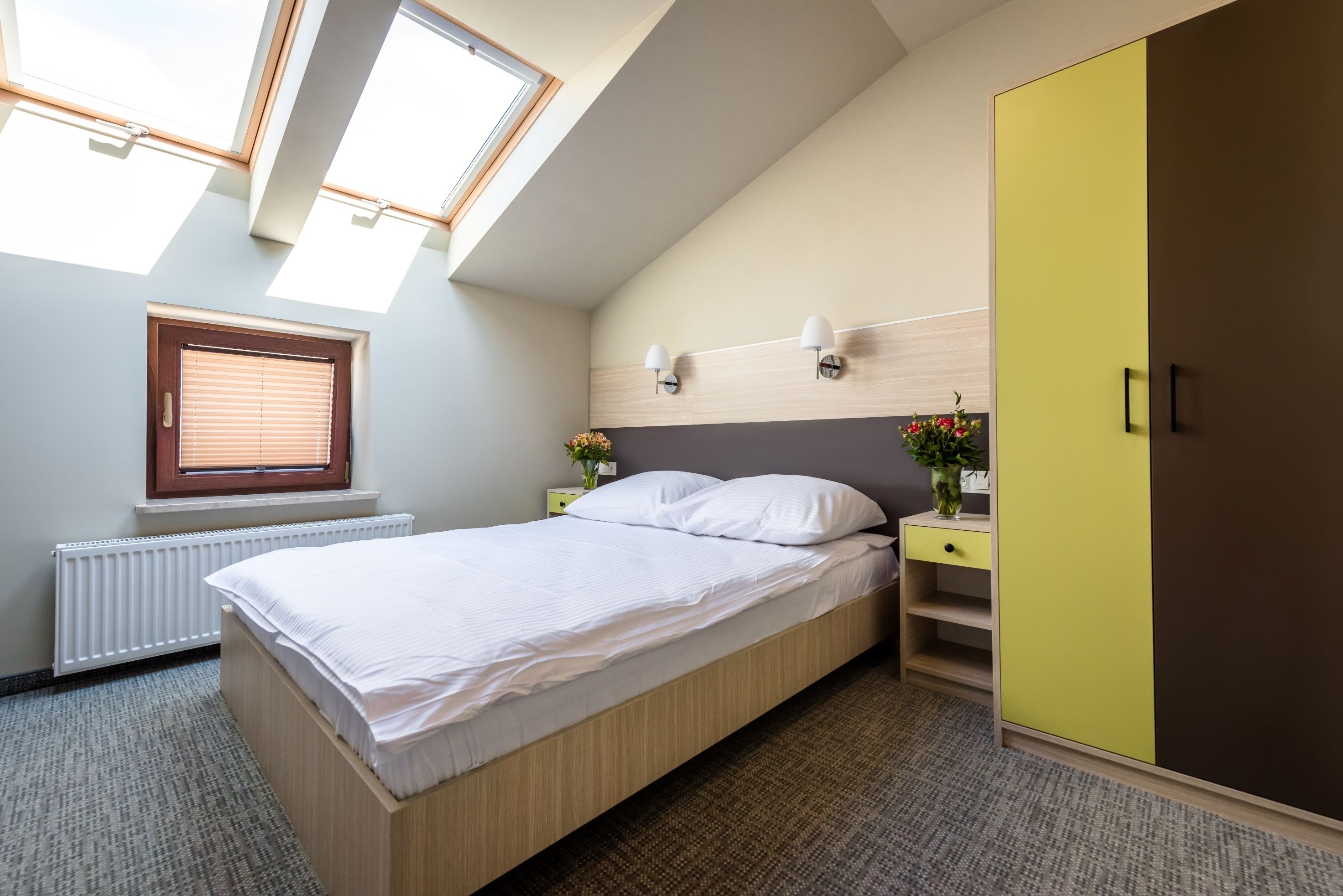 Ein Schlafzimmer mit Bett, einem Fenster mit Jalousien, einem Heizkörper, einem Nachttisch mit Lampe und einem Kleiderschrank.