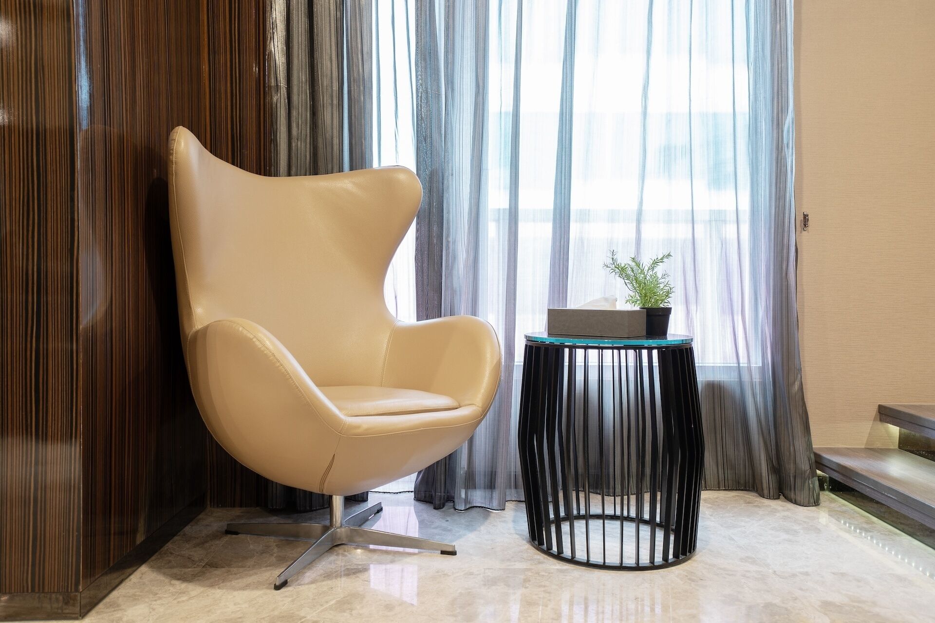 Una habitación moderna con una butaca de respaldo alto, una mesita redonda con una planta y cortinas traslúcidas.