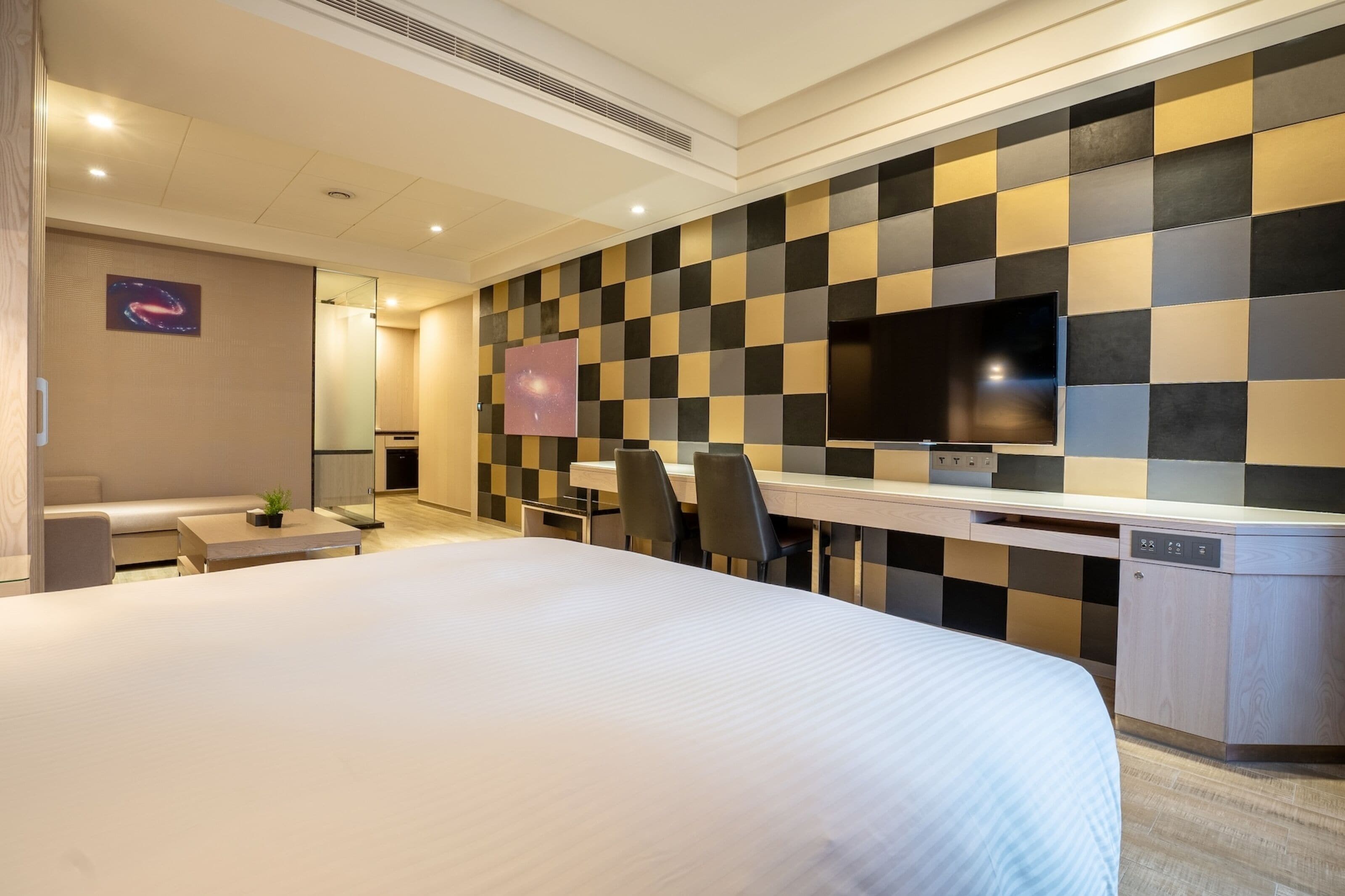 A hotel room with a large bed, a desk with a flat-screen TV, a seating area with a sofa and chairs, and a wall with a checkered pattern.