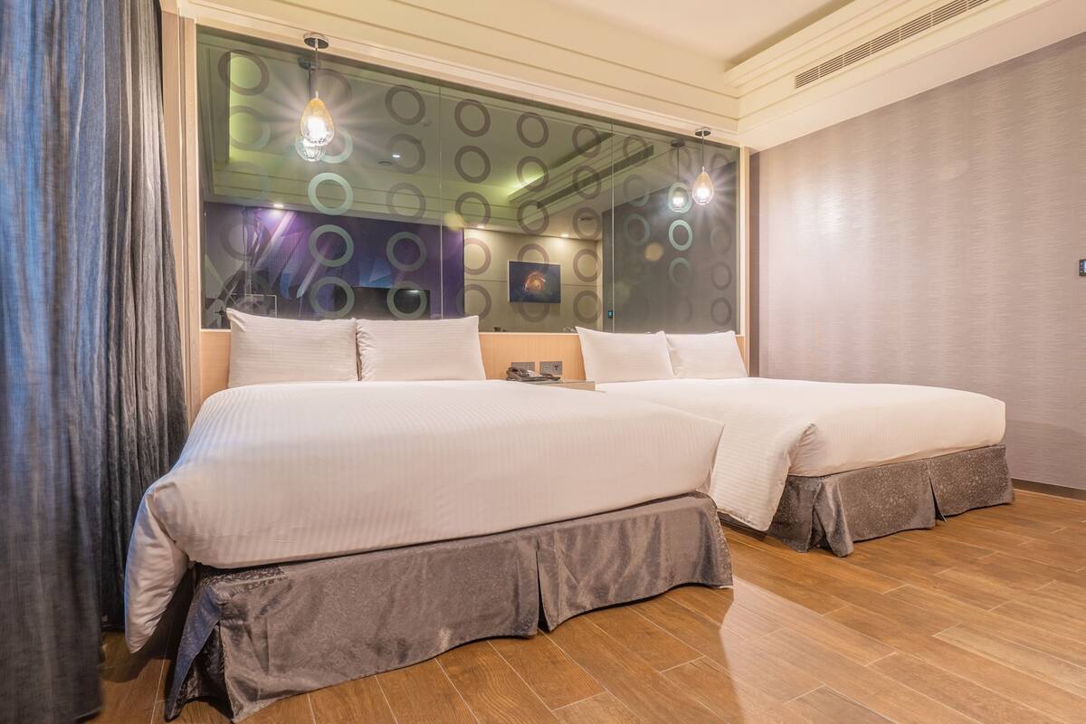 A hotel room with two beds, a large window with circular patterns, and a wooden floor.