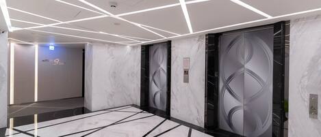 Un ascensor moderno con diseños geométricos en las puertas, suelo de mármol con líneas negras e iluminación lineal blanca en el techo.