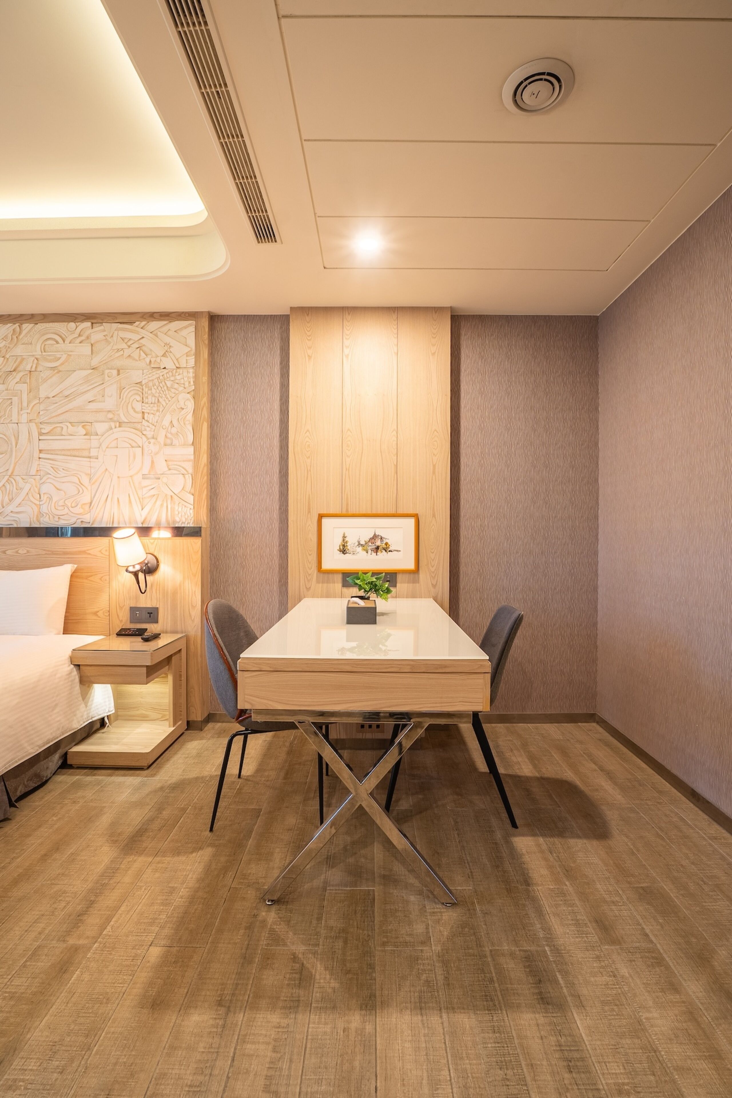 A hotel room with a bed, bedside table, lamp, wooden headboard, wall art, and a dining table with chairs.