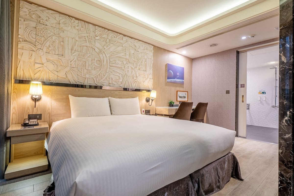 A hotel room with a large bed, a wooden headboard, a wall-mounted TV, a desk with a chair, and a bathroom visible through an open door.