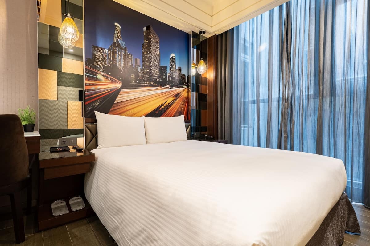 A hotel room with a large bed, a desk with a chair, a nightstand, and a cityscape mural on the wall.