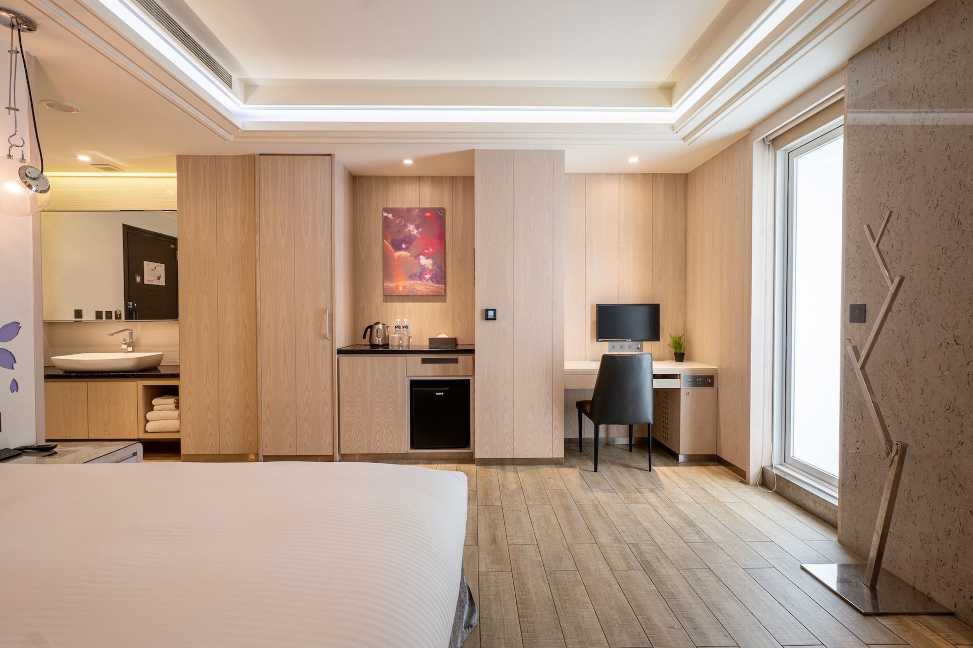 A modern hotel room with a large bed, a bathroom with a sink and mirror, a desk with a chair, and a TV.