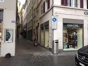 Une rue étroite avec un magasin GEOX, un panneau indiquant une zone piétonne et une publicité pour les montres TISSOT.