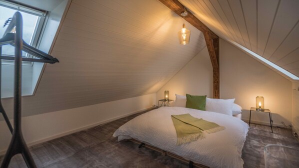 Une chambre confortable située sous les toits, avec un plafond en pente, un grand lit recouvert de linge de lit blanc, deux lampes de chevet et une lucarne.