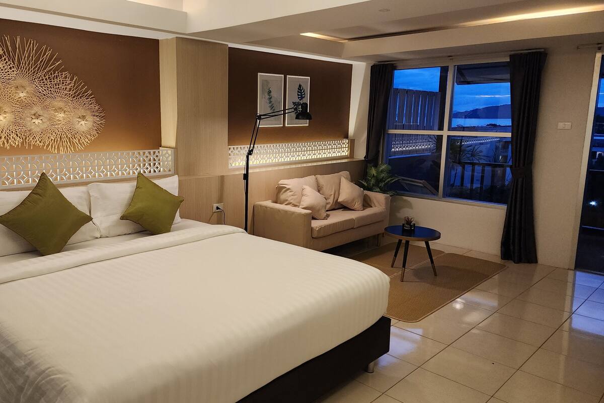 Une chambre d’hôtel moderne avec un grand lit, un canapé, une petite table et une vue sur l’extérieur par une fenêtre.