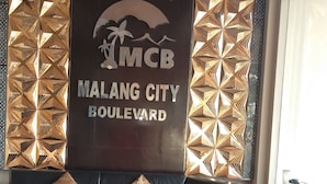 Un salon avec un canapé d’angle noir, des coussins décoratifs et un mur orné d’une enseigne indiquant : « Home Stay MCB MALANG CITY BOULEVARD ».