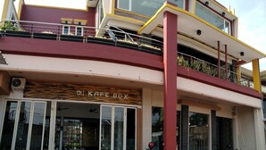 Un bâtiment à deux étages abritant un café appelé « KAFE BOX » au rez-de-chaussée. Des espaces extérieurs avec des tables et des chaises sont aménagés pour accueillir les clients.