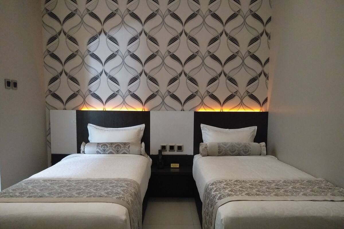 A hotel room with two beds, a patterned wallpaper, and a nightstand with a lamp.