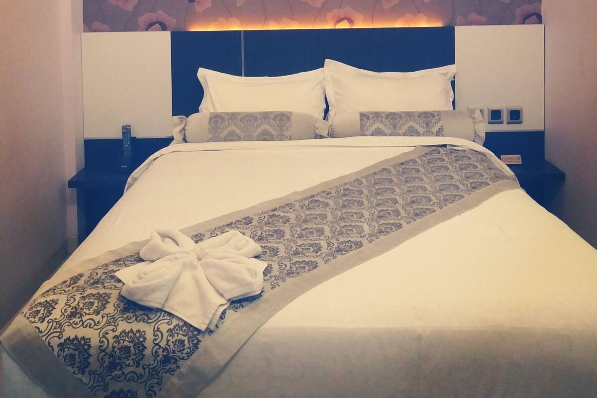 A hotel room with a large bed, a headboard with a floral pattern, and a nightstand with a lamp.